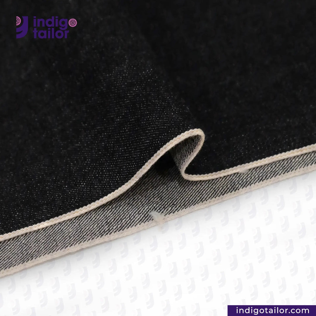 Close-up of BLACK SLUBBY SELVEDGE DENIM fabric with beige edge on a white background, featuring Indigotailor logo.