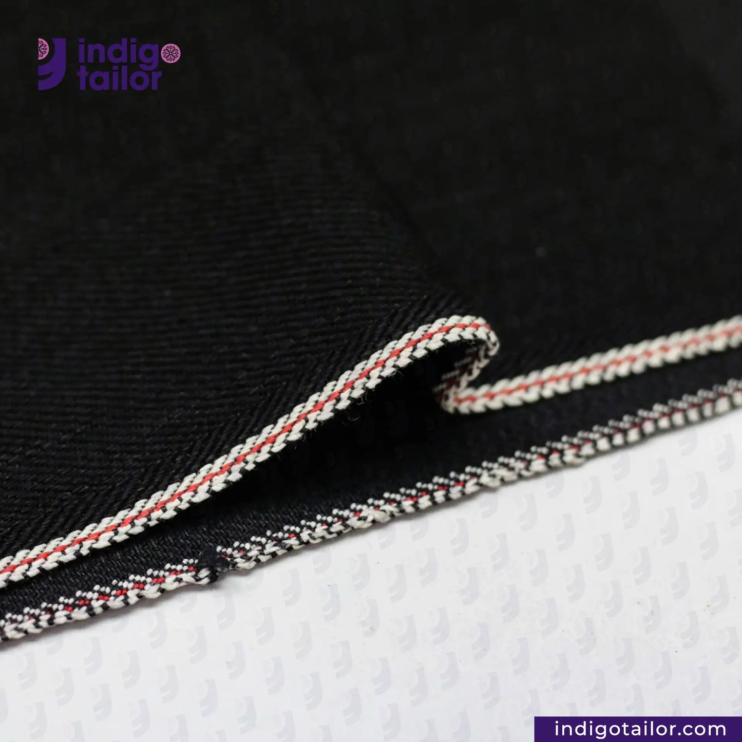 Close-up of BLACK X BLACK SELVEDGE DENIM fabric with white and red trim, branded 'Indigotailor'.