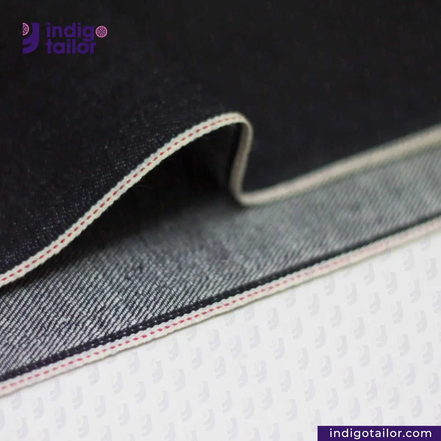 Close-up of DEEP INDIGO DOTTED BRONZE SELVEDGE denim fabric with visible stitching on a white background, featuring the 'indigotailor' brand.