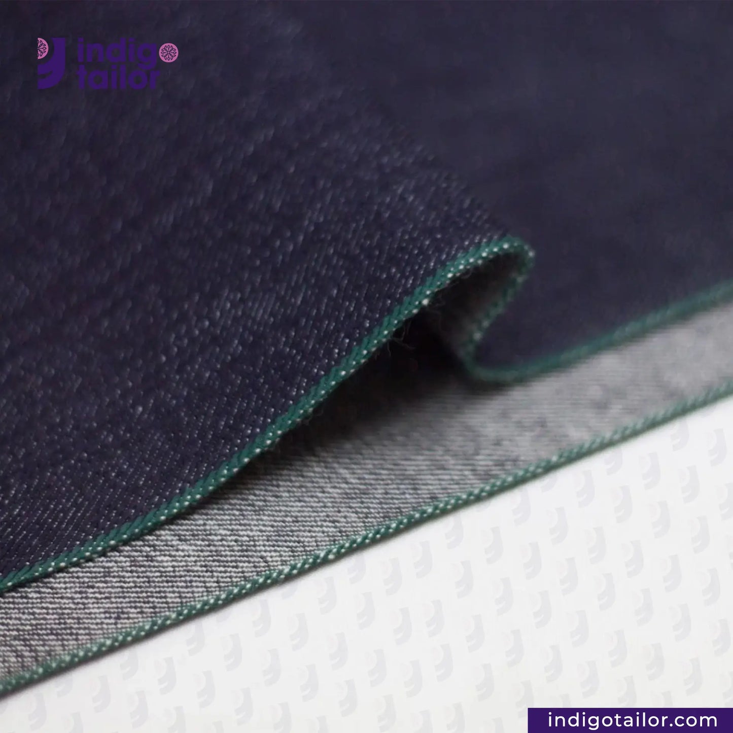 Close-up of DEEP INDIGO FOREST GREEN SELVEDGE DENIM fabric with green stitching on a white background, featuring 'Indigo Tailor' branding.