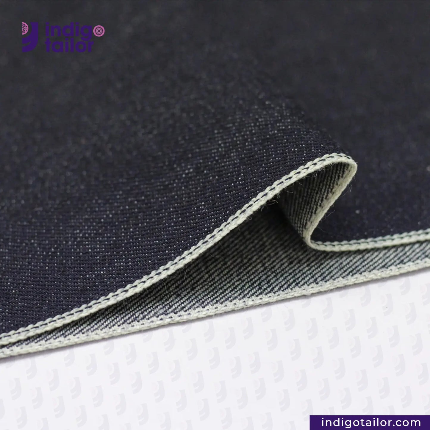 Close-up of DEEP INDIGO GREENCAST SELVEDGE DENIM fabric with visible stitching on a white background, featuring the 'Indigo Tailor' brand.