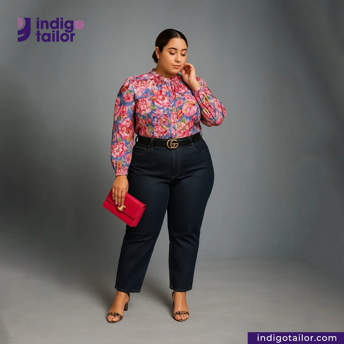 Woman wearing a floral blouse and DEEP INDIGO GREENCAST SELVEDGE DENIM pants with a red clutch, standing against a gray background with 'Indigotailor' branding.