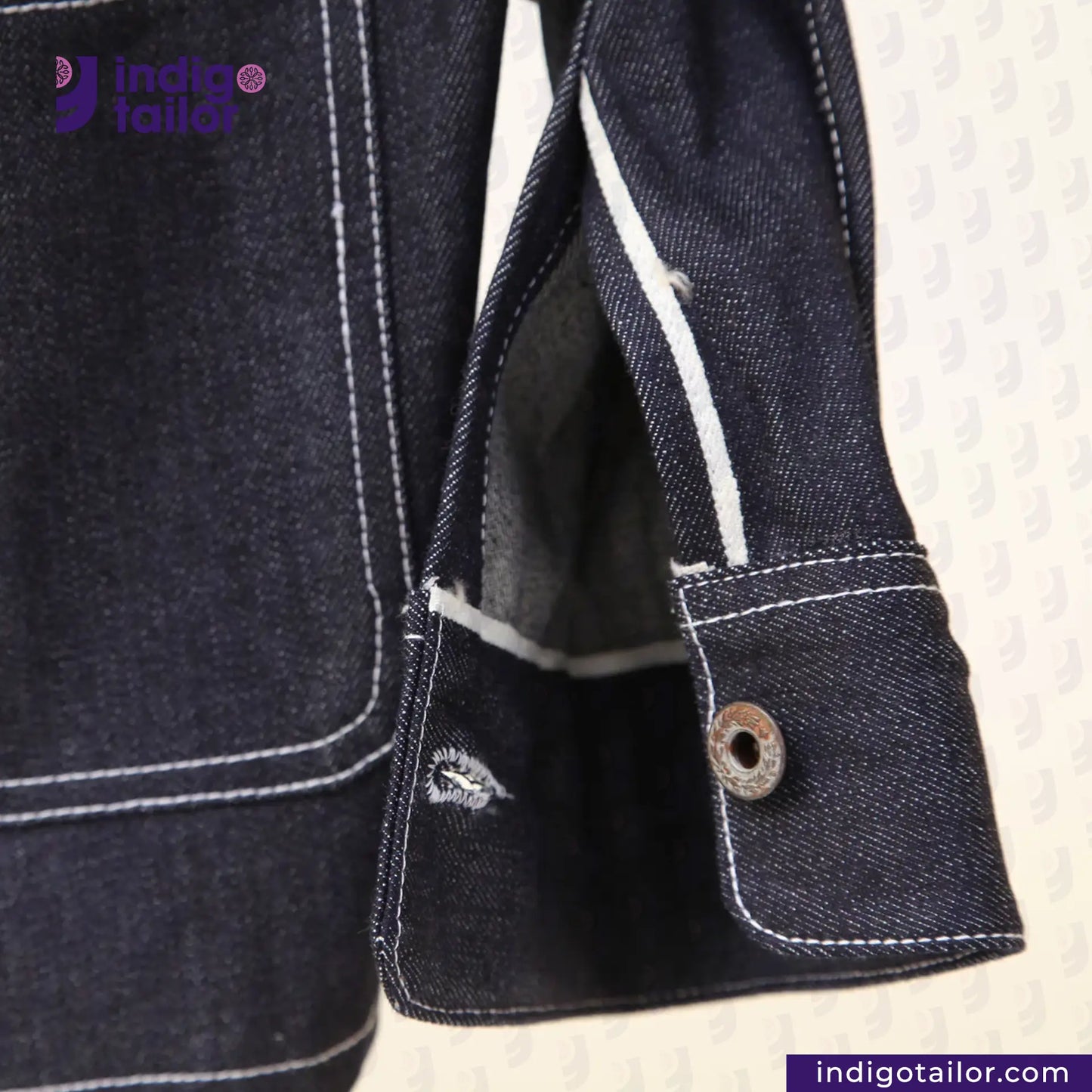 Close-up of a DENIM CHORECOAT jacket with white stitching on a textured beige background, featuring the Indigo Tailor logo.