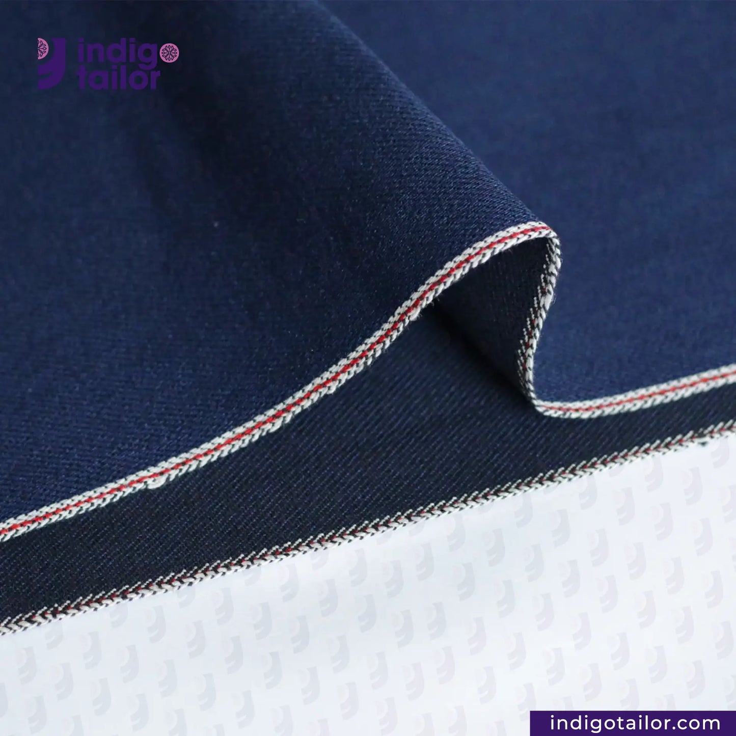 Close-up of DOUBLE DIP INDIGO SELVEDGE DENIM fabric with red and white trim, featuring 'Indigotailor' branding.