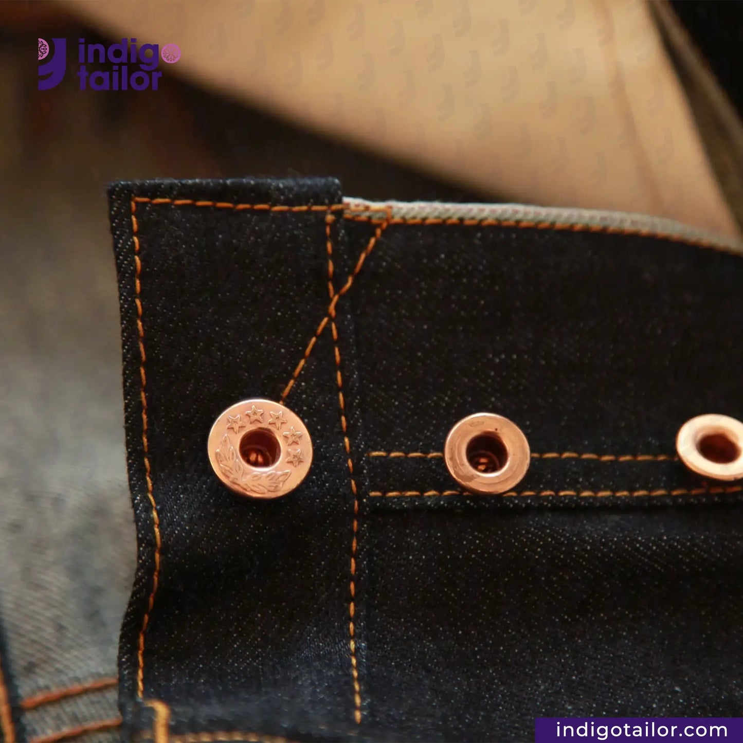 Close-up of black denim fabric with copper buttons and stitching, featuring 'indigotailor' branding.