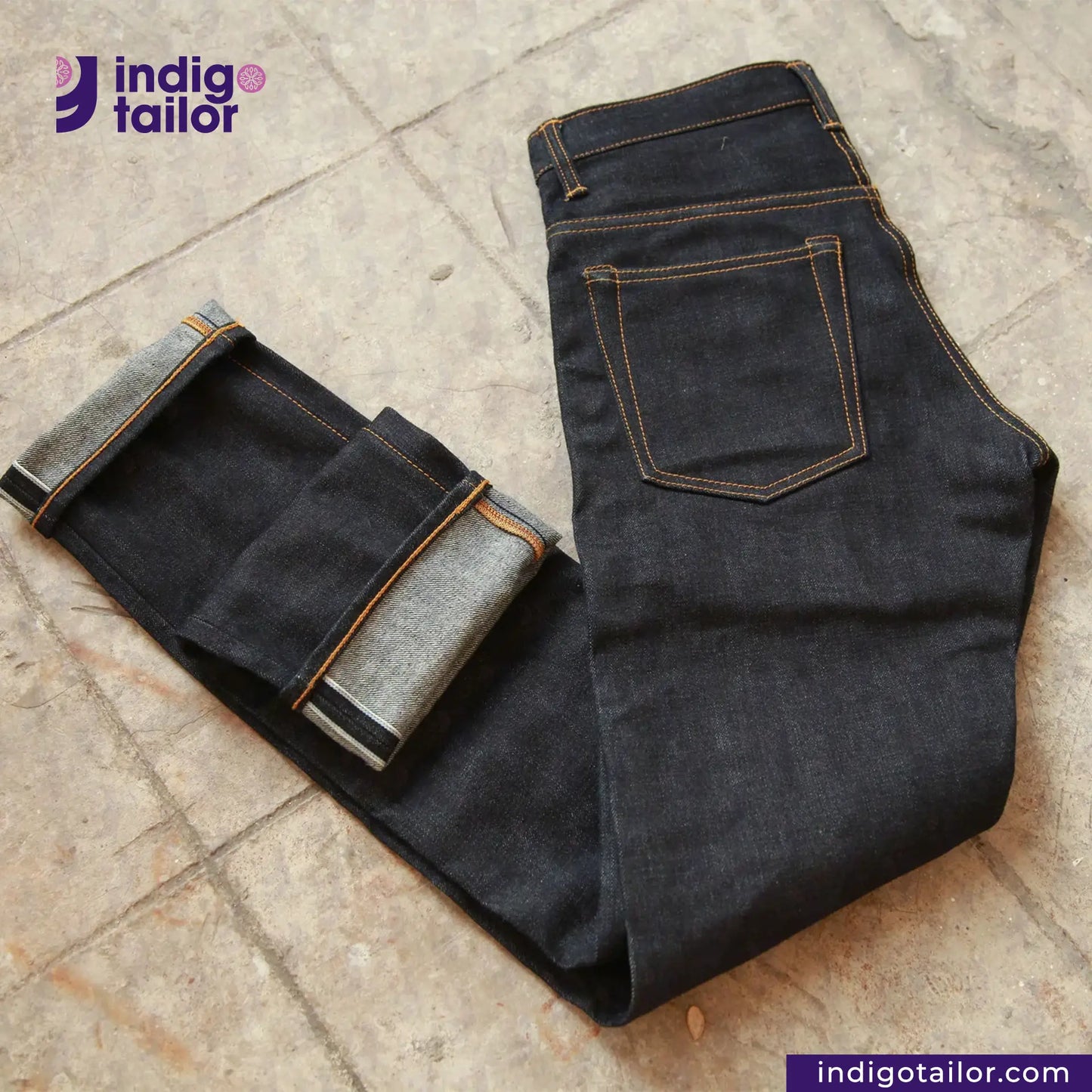 Unfolded and folded dark blue jeans on a textured surface with 'indigotailor' branding.