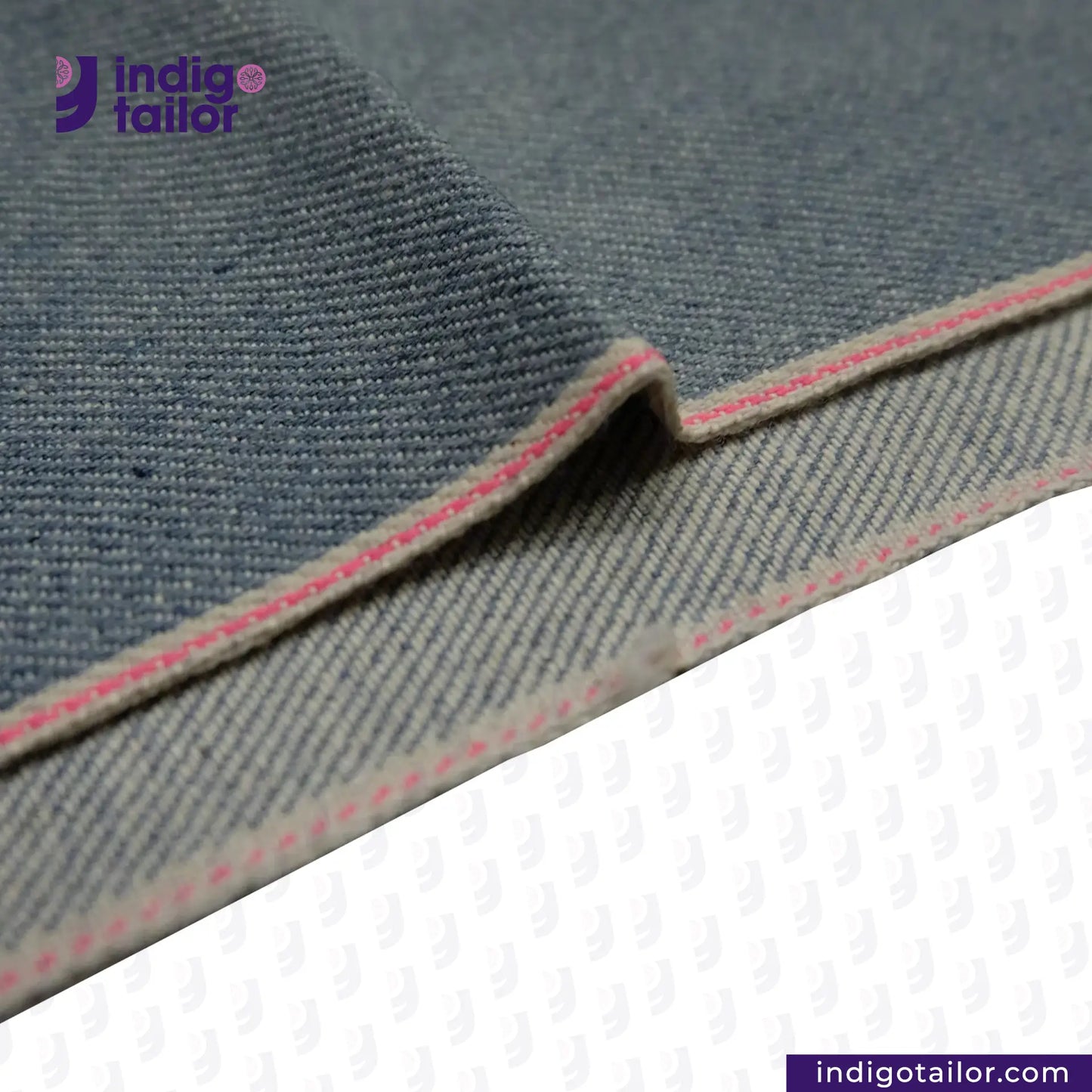 Close-up of ICE BLUE SELVEDGE DENIM fabric with pink stitching on a white background, featuring the Indigo tailor logo.