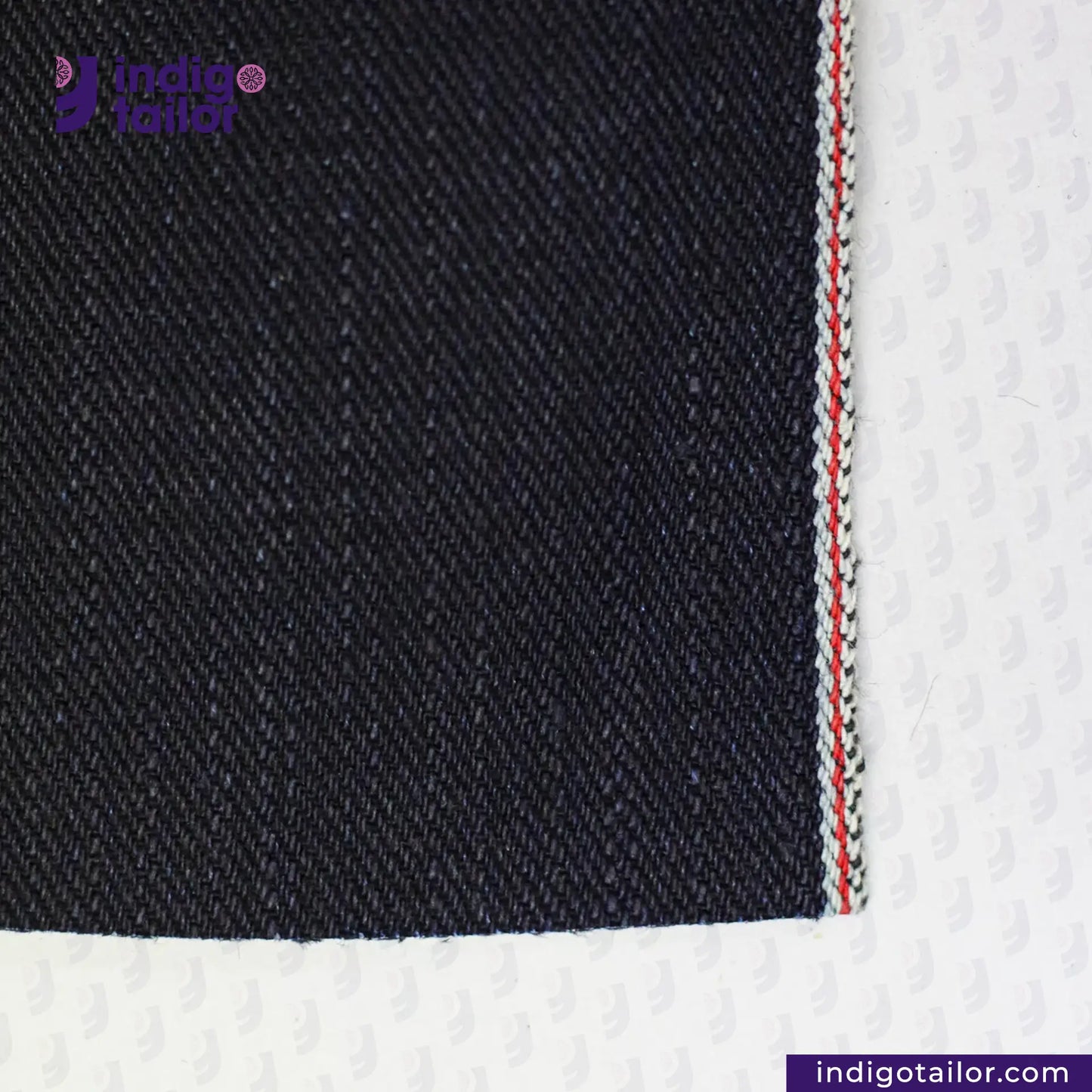 Close-up of INDIGO X BLACK DENIM fabric with a white border and red stitching, featuring the Indigo Tailor logo.