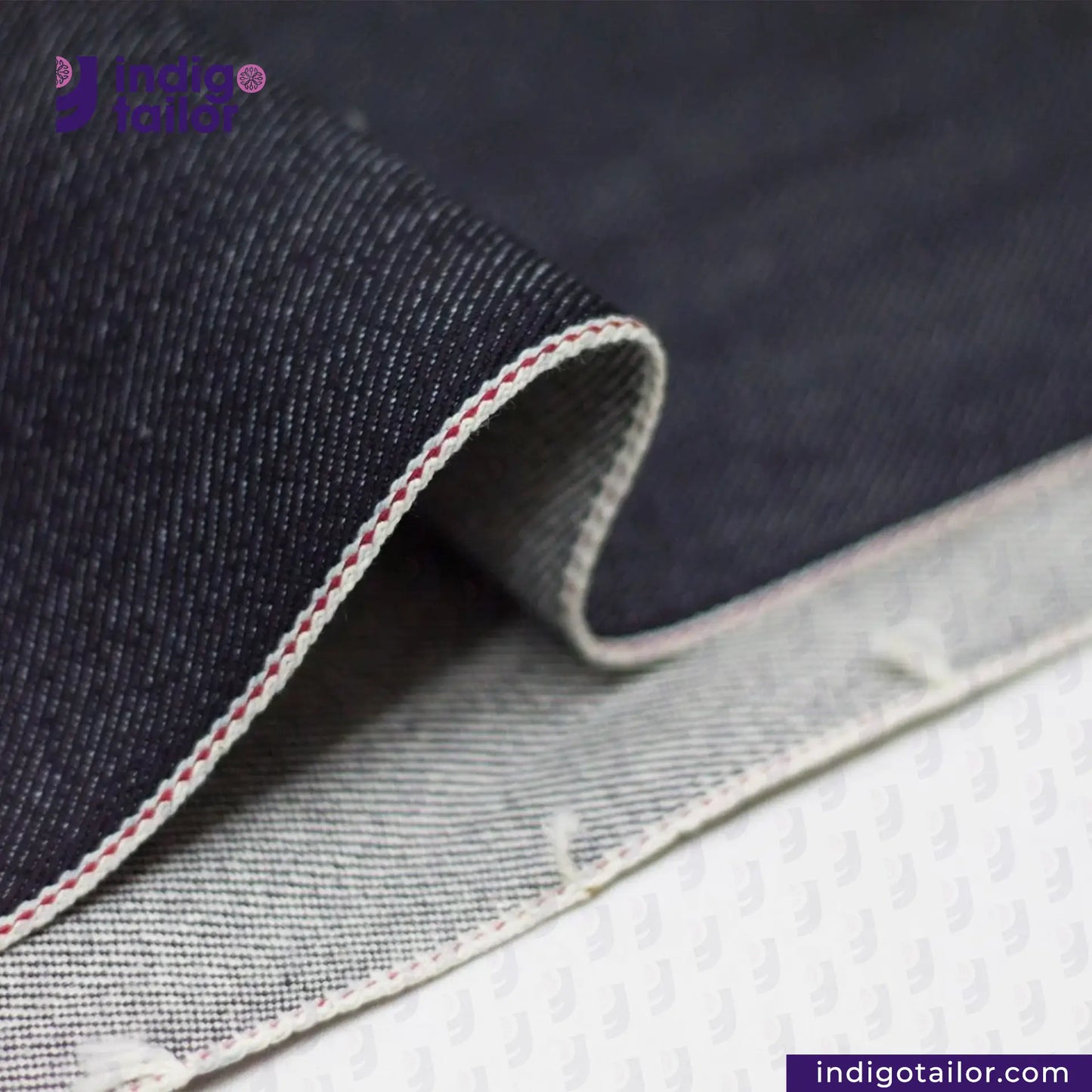 Close-up of IRONMAN HEAVYWEIGHT SELVEDGE DENIM fabric with visible stitching on a textured surface, featuring the Indigo Tailor logo.