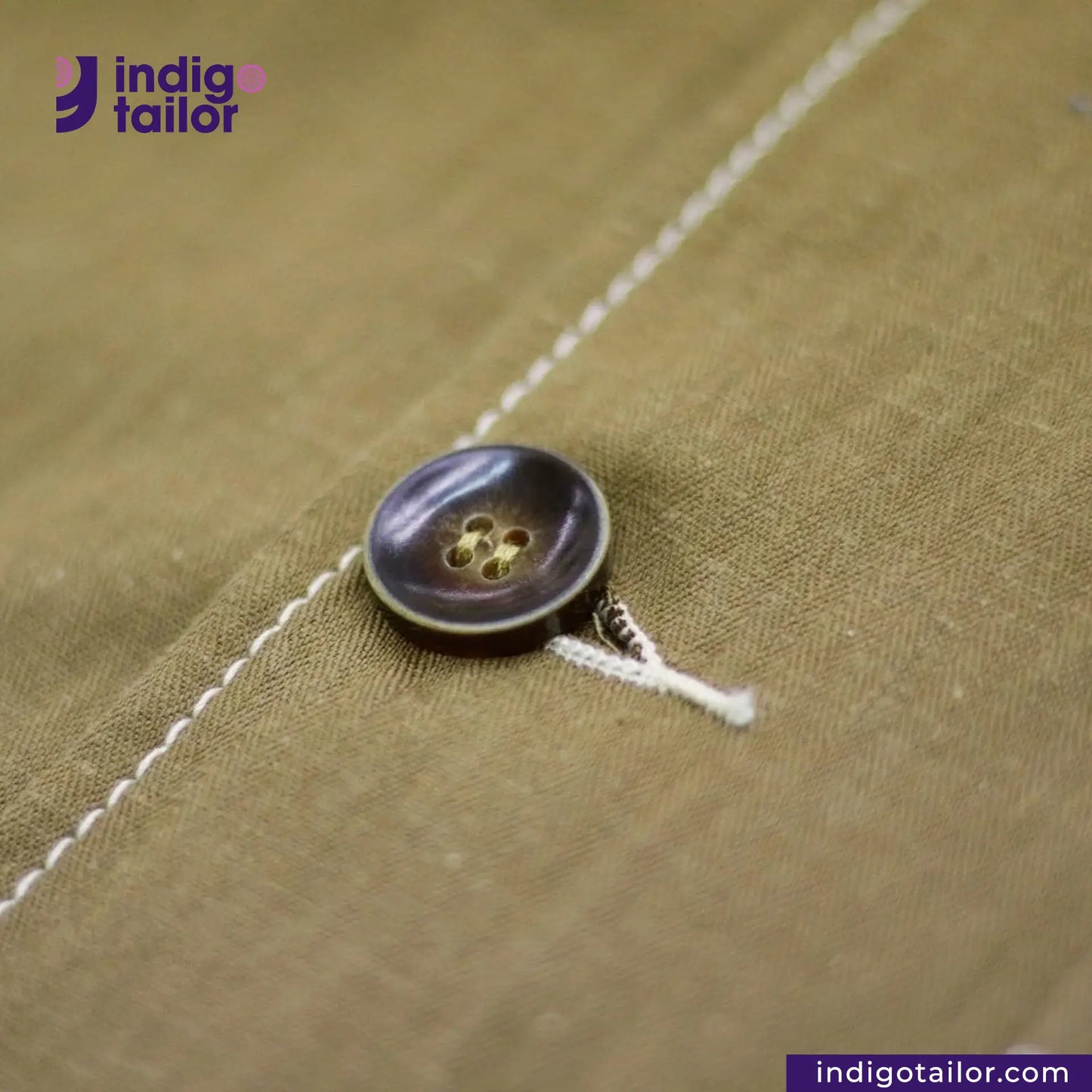 Close-up of a button on KHAKI HERRINGBONE COVERALL JACKET with 'indig tailor' branding.