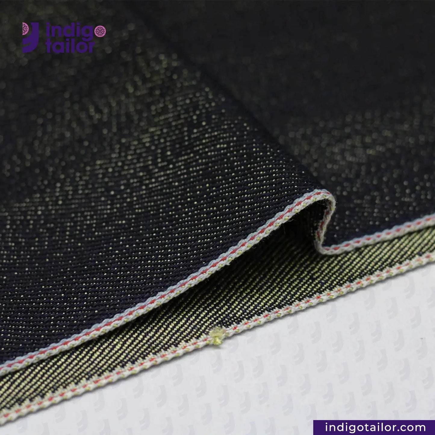 Close-up of METALLIC GOLD WEFT SELVEDGE DENIM fabric with visible stitching on a white background, featuring the 'Indigotailor' brand.