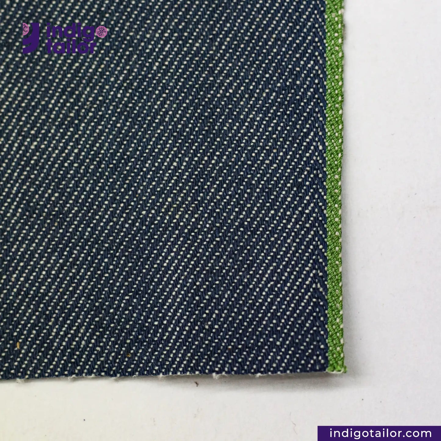 Close-up of OCEAN BLUE COTTON HEMP BLEND SELVEDGE DENIM fabric with a green edge on a white background, featuring the Indigo Tailor logo.