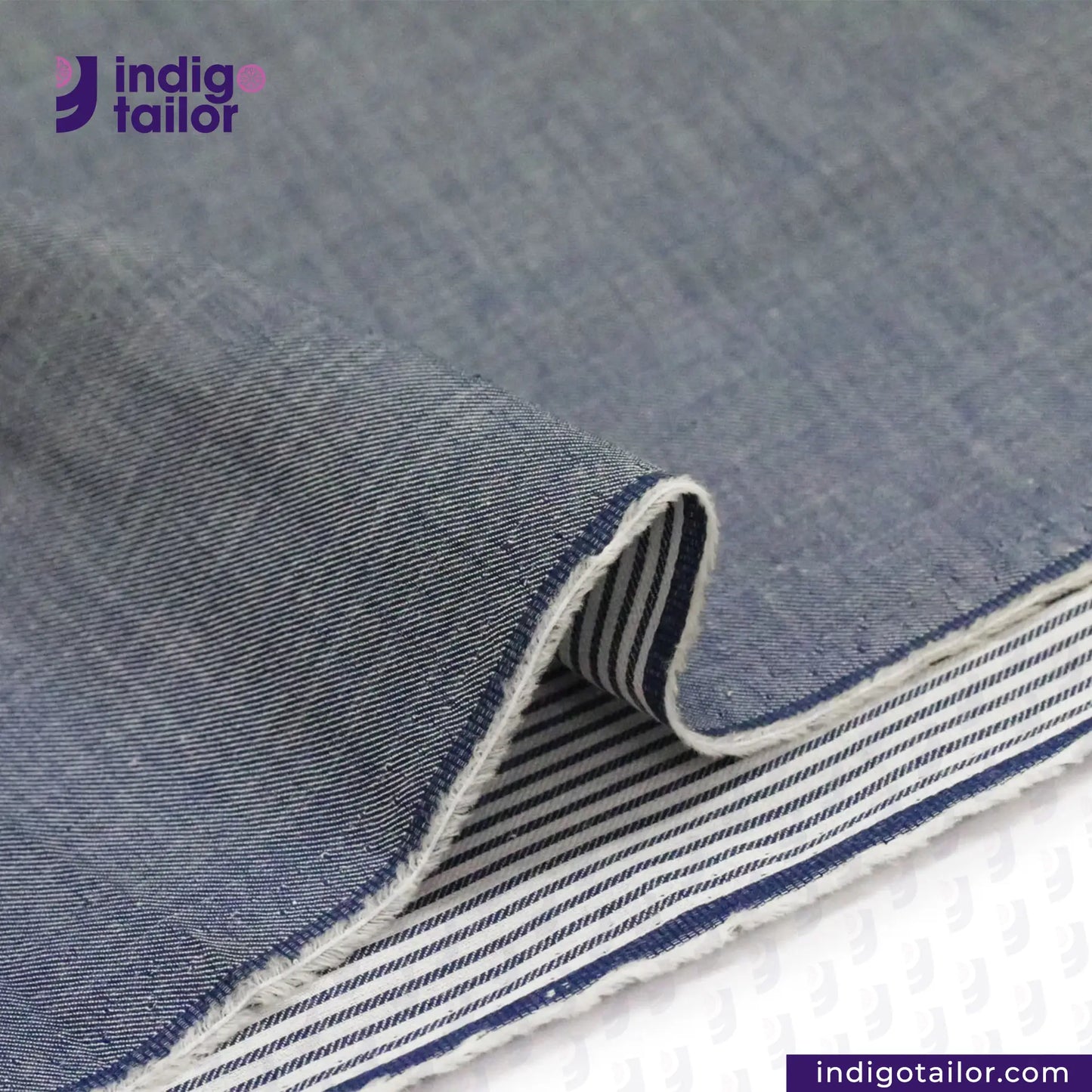 Close-up of REVERSIBLE CHAMBRAY BLUE GREY WITH SUBTLE HICKORY STRIPES fabric with 'indigotailor' branding.