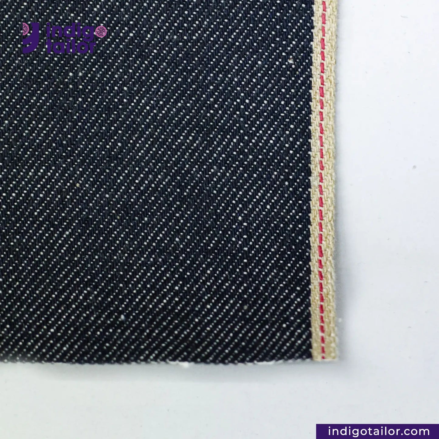 Close-up of RIGID INDIGO SELVEDGE DENIM fabric with a red and white striped edge on a white background, featuring the Indigo Tailor logo.