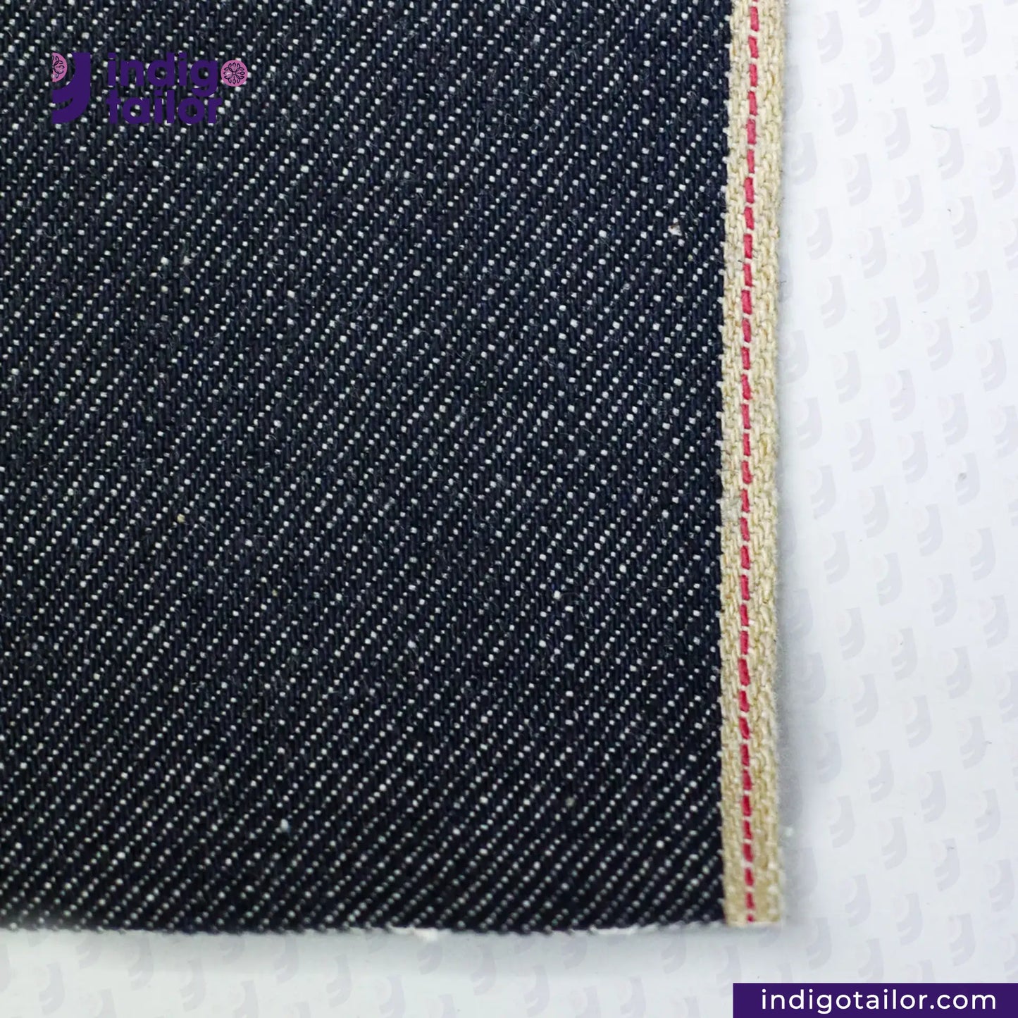Close-up of RIGID INDIGO SELVEDGE DENIM fabric with a visible seam on a white background, featuring the Indigotailor.com logo.