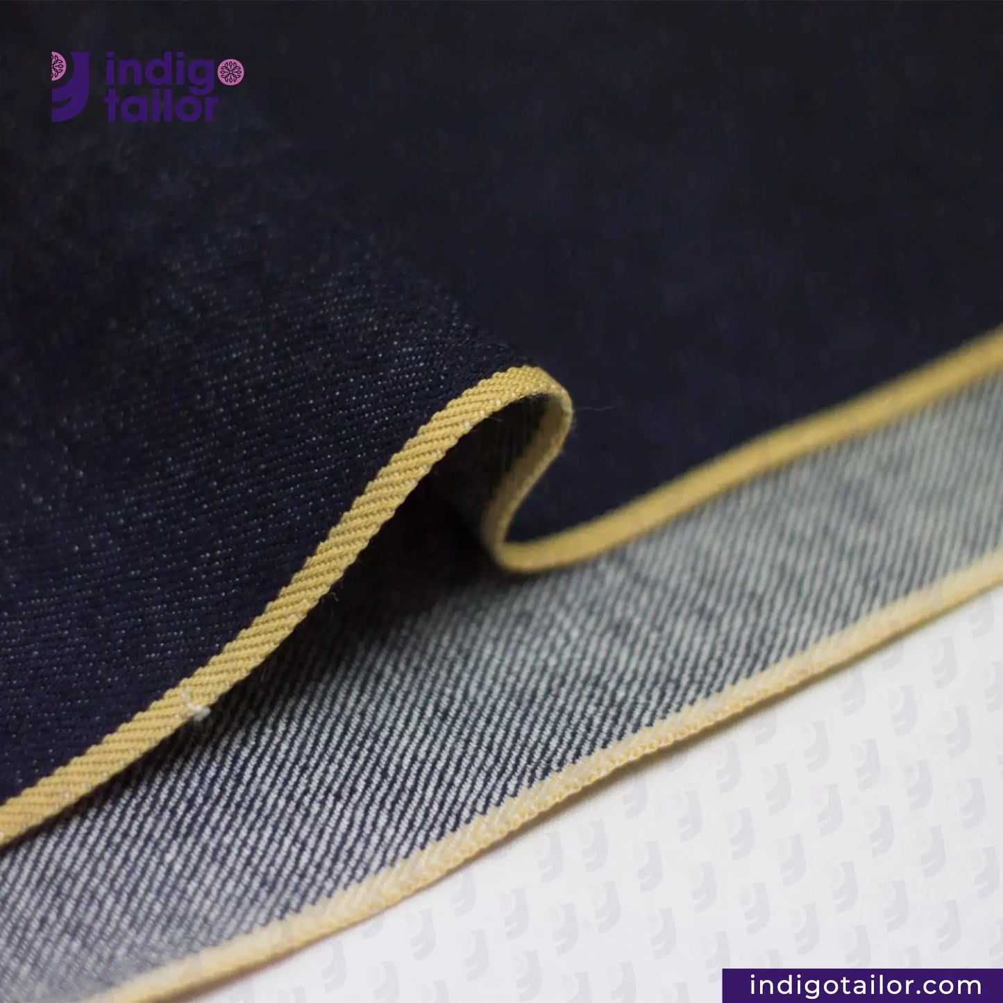 Close-up of SATURATED INDIGO GOLDEN SELVEDGE fabric with beige stitching on a textured surface, featuring 'Indigotailor' branding.