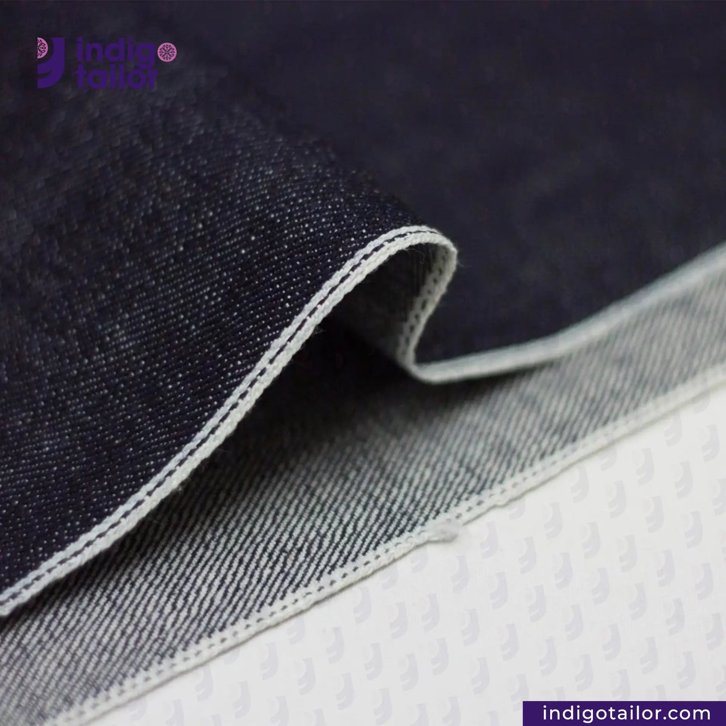 Close-up of SLUBBY INDIGO SELVEDGE DENIM fabric with visible stitching, brand 'Indigo Tailor' in the corner.