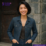 Woman wearing a SPACE GREY DENIM jacket standing against a stone wall with 'Indigo Tailor' branding.