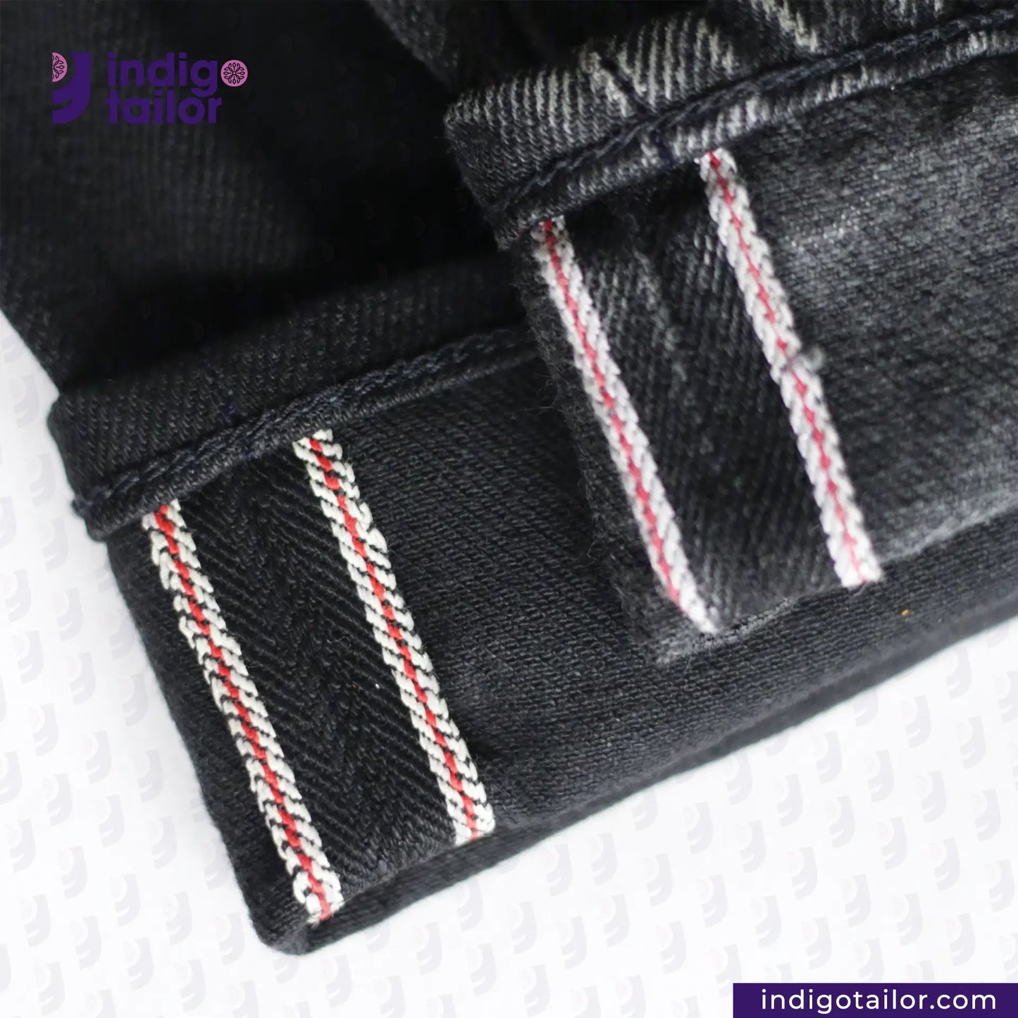 Close-up of SUPER BLACK DENIM jeans with white and red stitching on a white background, featuring the Indigotailor logo.