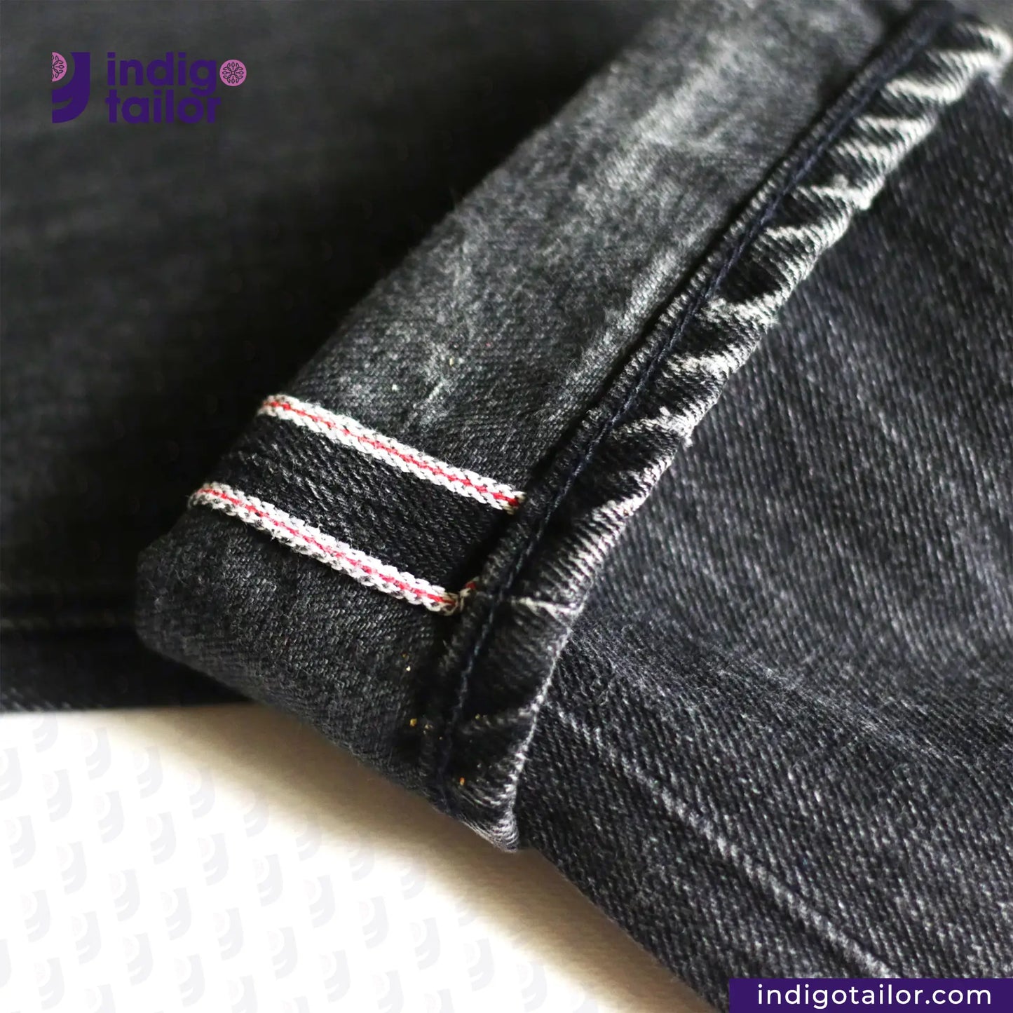 Close-up of SUPER BLACK DENIM denim fabric with white stitching on a white background, featuring the Indigo Tailor logo.