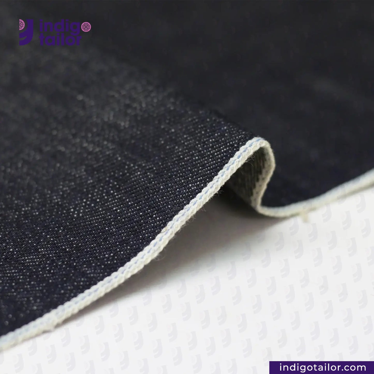 Close-up of TATE OCHI DOUBLE RING SPUN SELVEDGE DENIM fabric with visible stitching on a white background, featuring the Indigotailor logo.