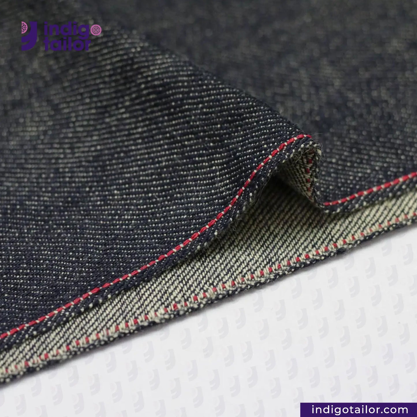 Close-up of TATE OCHI DOUBLE RING SPUN SELVEDGE DENIM fabric with red stitching on a white background, featuring the Indigo Tailor logo.