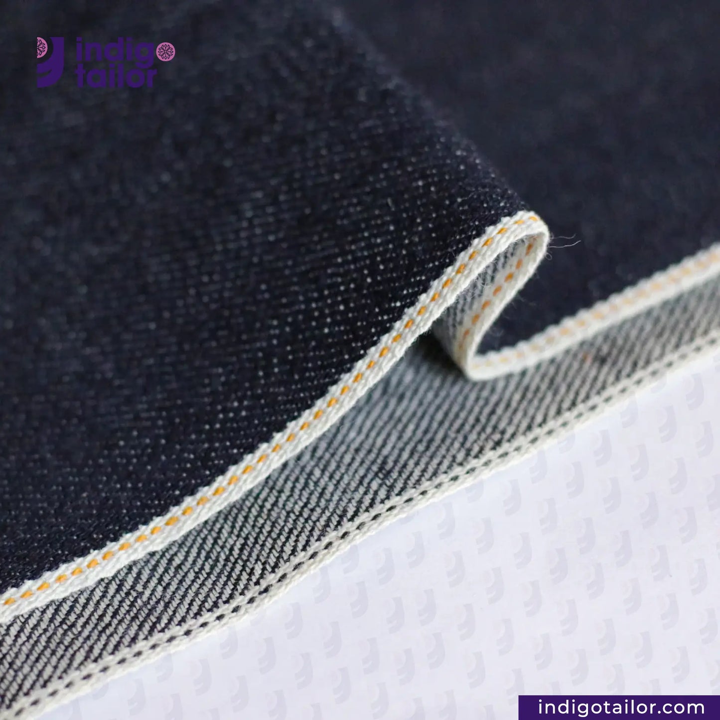 Close-up of TWO TONE SELVEDGE PAPER WEIGHT DENIM fabric with visible stitching on a white background, featuring the Indigotailor logo.
