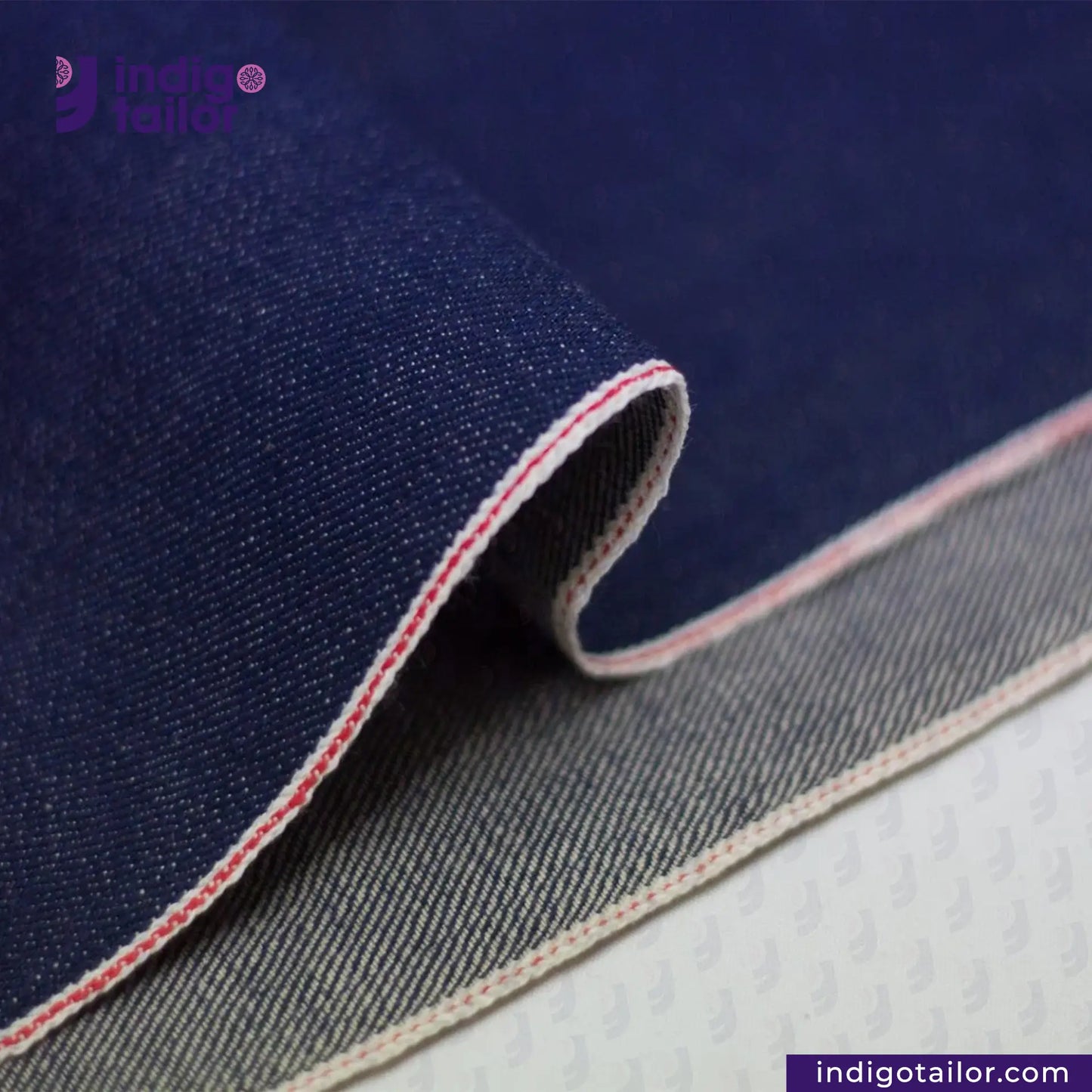 Close-up of VINTAGE BLUE SELVEDGE DENIM fabric with red stitching on a blue background, featuring the Indigotailor logo.