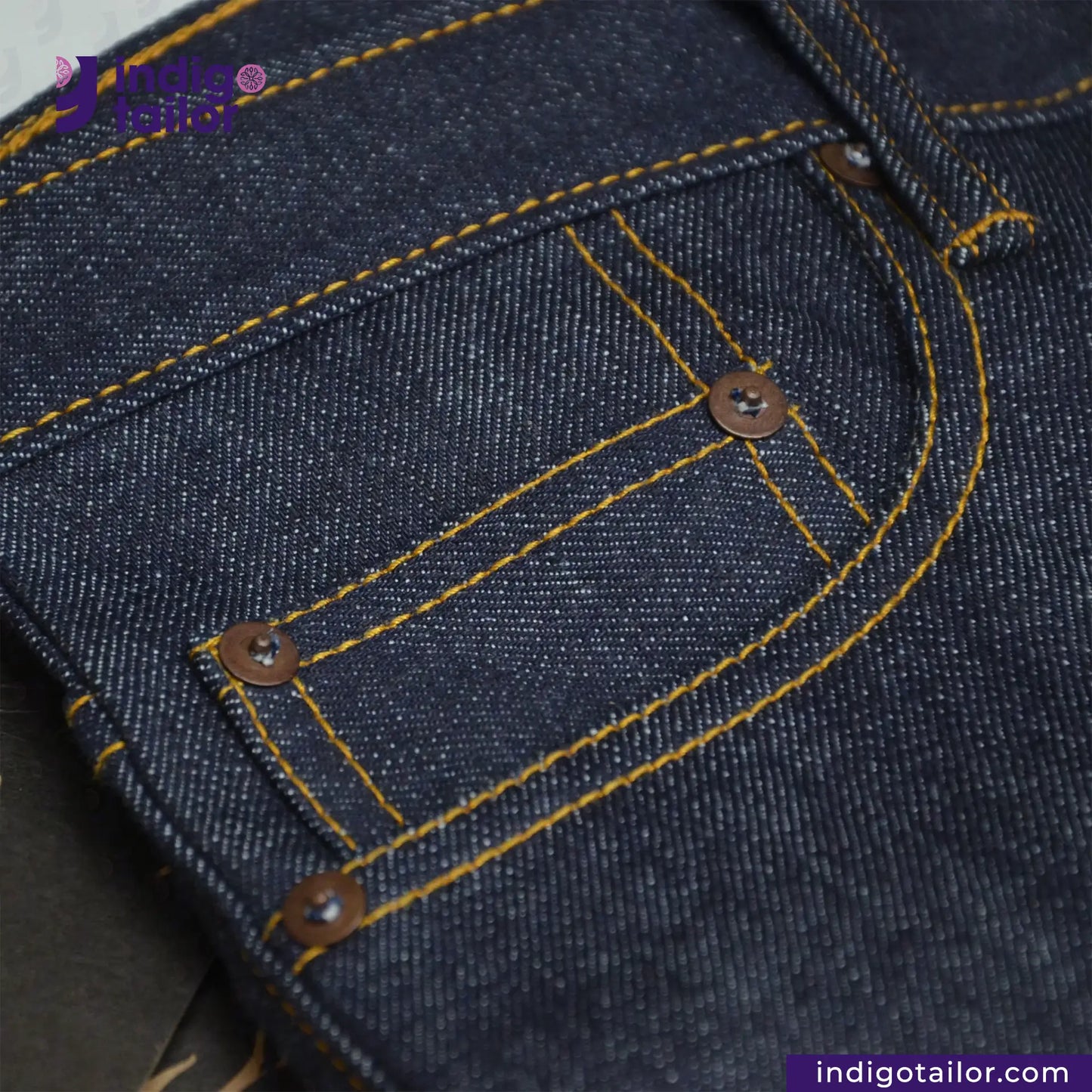 Close-up of VINTAGE SLUB DENIM jeans pocket area with yellow stitching, featuring 'Indigo Tailor' branding.