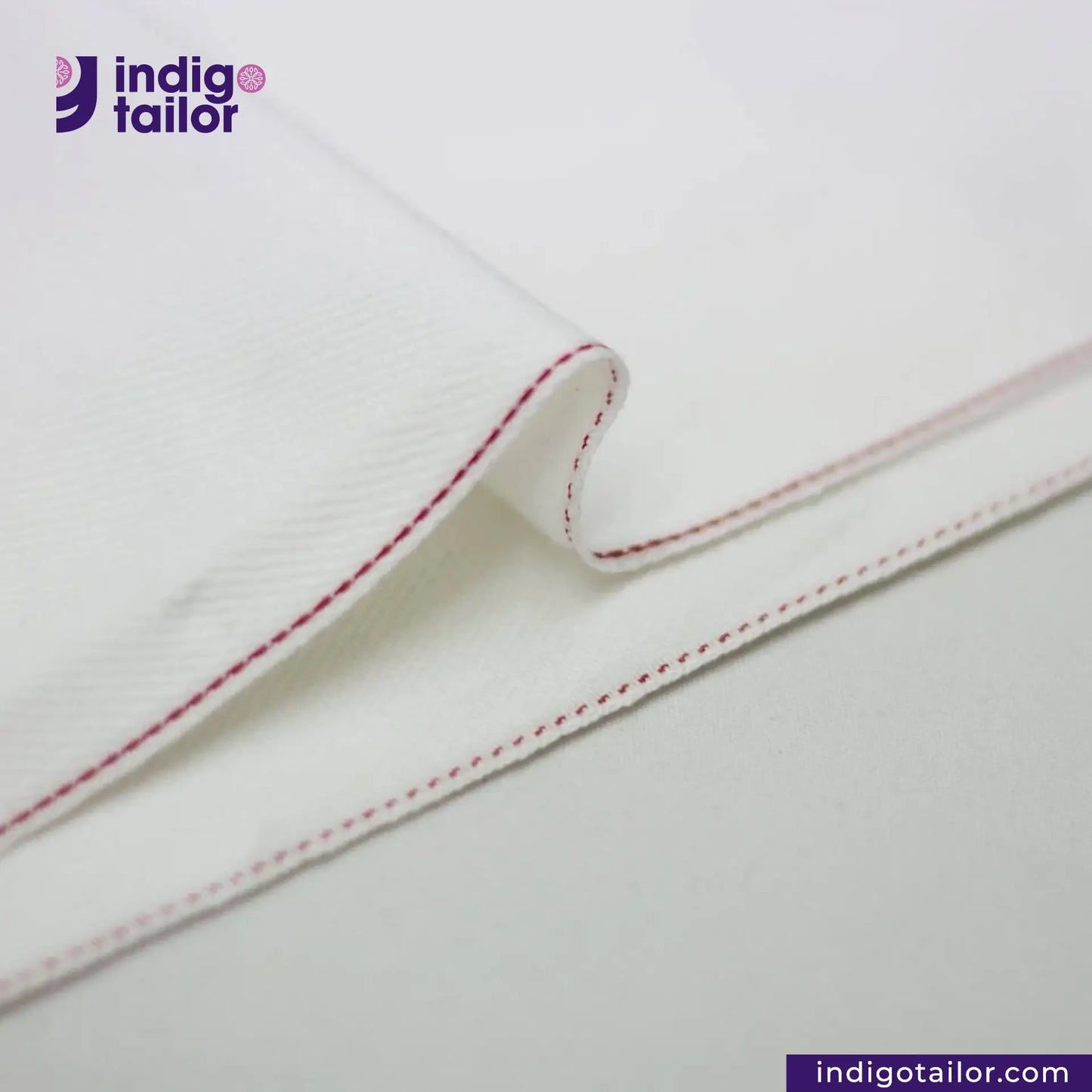 Close-up of WHITE DENIM DOTTED RED SELVEDGE fabric with red stitching on a light gray background, featuring the Indigotailor logo.