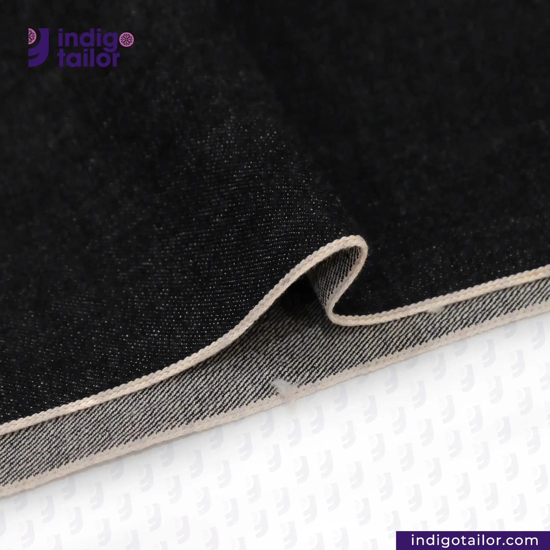 Close-up of BLACK SLUBBY SELVEDGE DENIM fabric with beige edge on a white background, featuring Indigotailor logo.