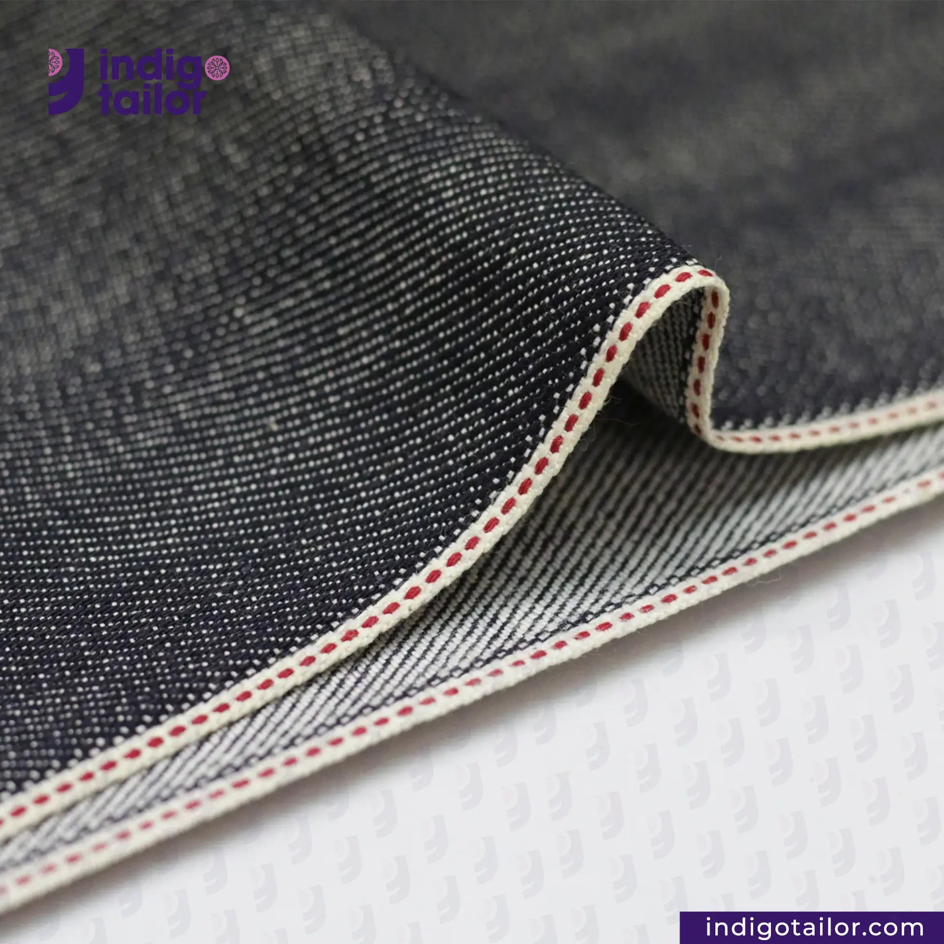 Close-up of BLUE GRAY SELVEDGE DENIM fabric with red stitching on a white background, featuring the Indigo Tailor logo.