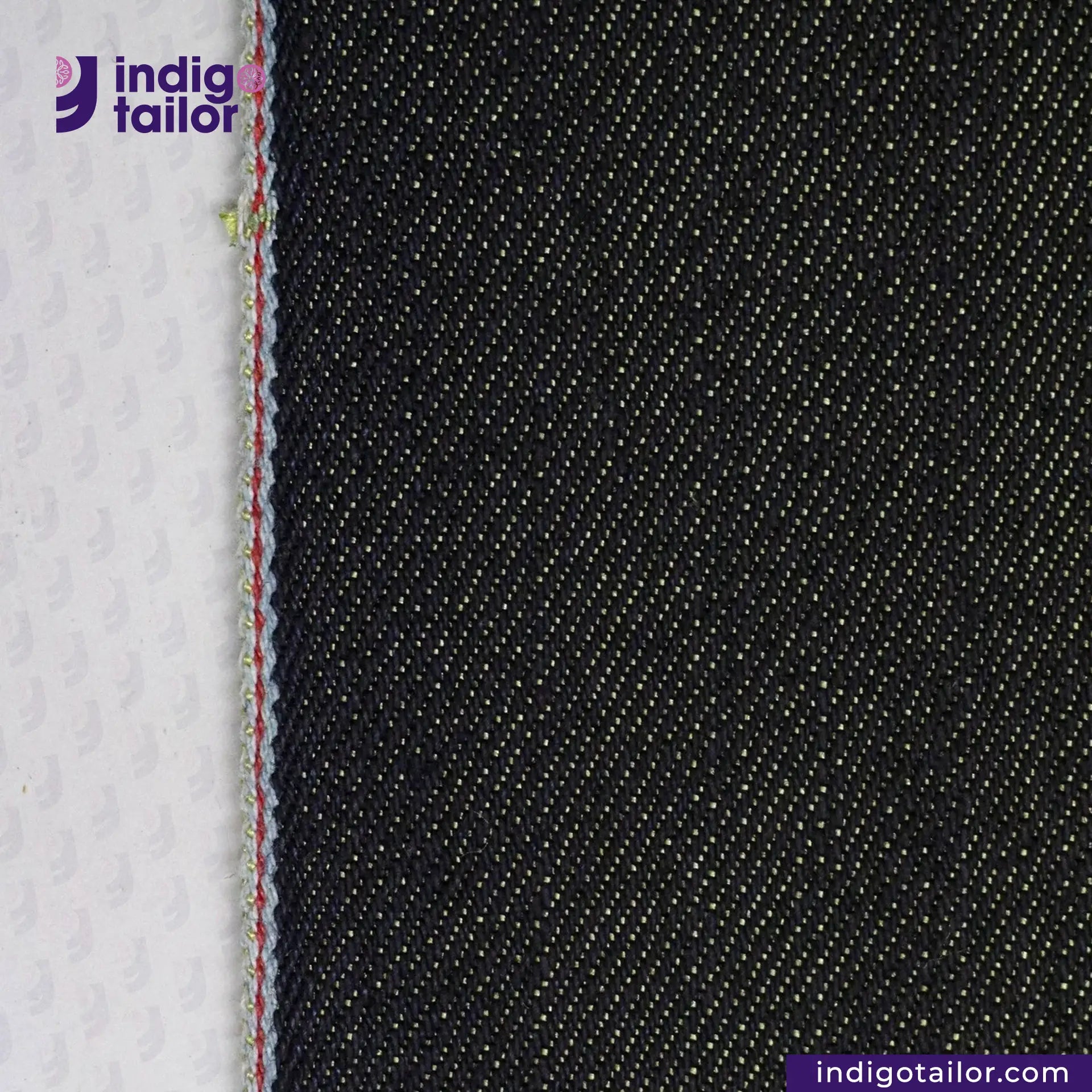 Close-up of black denim fabric with a white background, featuring the Indigotailor logo. BULLION GOLD SELVEDGE DENIM