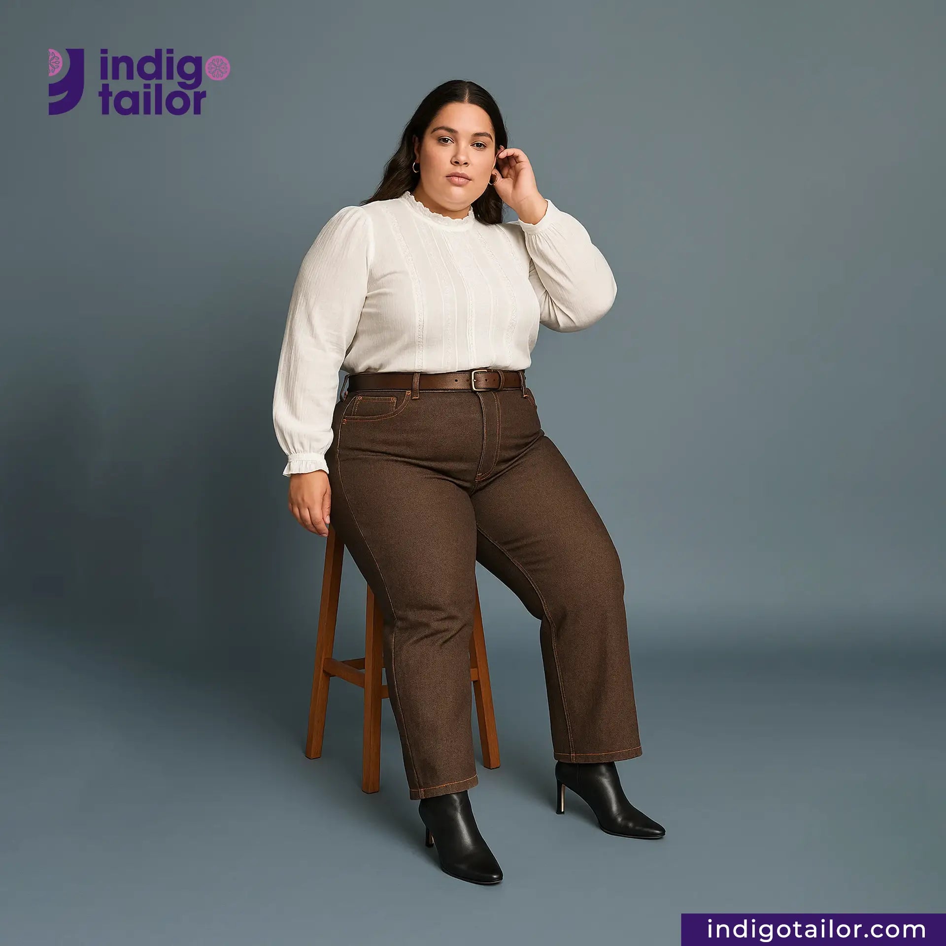 Woman wearing a white blouse and brown pants sitting on a stool with Indigotailor logo in the corner. Brown SOFT SLUB SELVEDGE DENIM