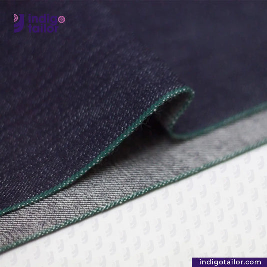 Close-up of DEEP INDIGO FOREST GREEN SELVEDGE DENIM fabric with green stitching on a white background, featuring 'Indigo Tailor' branding.