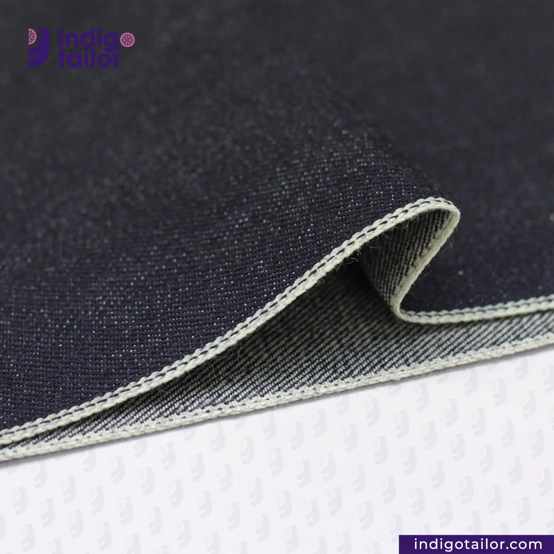 Close-up of DEEP INDIGO GREENCAST SELVEDGE DENIM fabric with visible stitching on a white background, featuring the 'Indigo Tailor' brand.