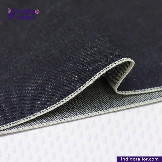 Close-up of DEEP INDIGO GREENCAST SELVEDGE DENIM fabric with visible stitching on a white background, featuring the 'Indigo Tailor' brand.