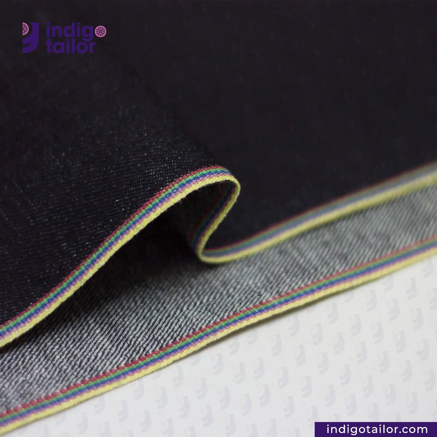 Close-up of DEEP INDIGO RAINBOW STRETCH SELVEDGE DENIM fabric with a colorful stripe on a textured surface, featuring the Indigotailor logo.