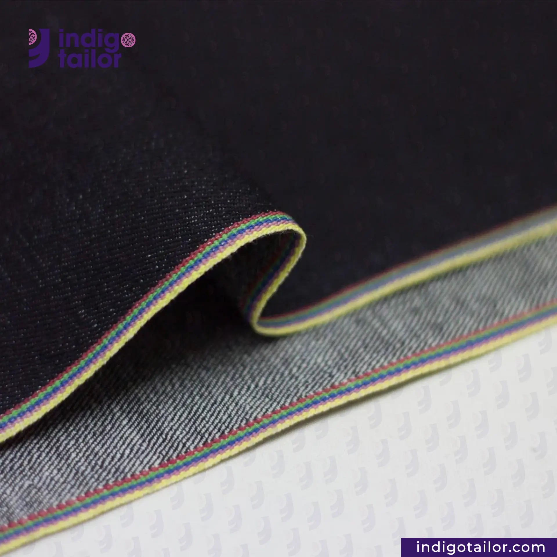 Close-up of DEEP INDIGO RAINBOW STRETCH SELVEDGE DENIM fabric with a colorful stripe on a textured surface, featuring the Indigotailor logo.