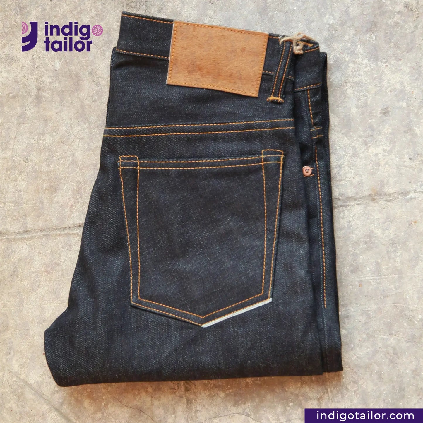 Folded pair of jeans with a brown patch on a textured surface, featuring the Indigotailor logo.