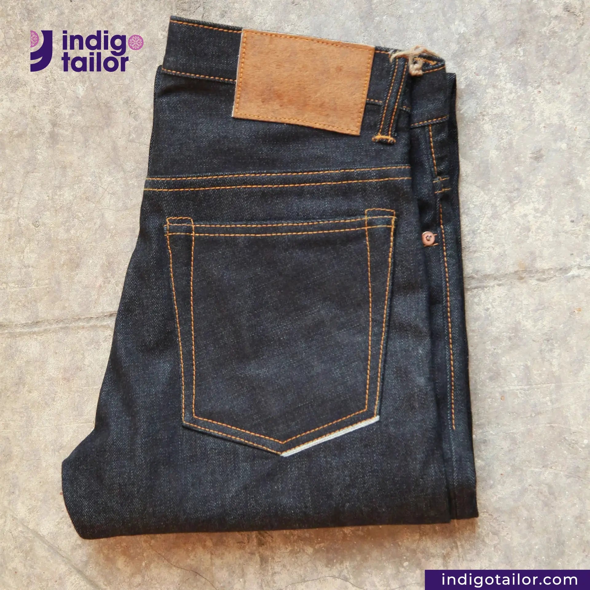 Folded pair of jeans with a brown patch on a textured surface, featuring the Indigotailor logo.