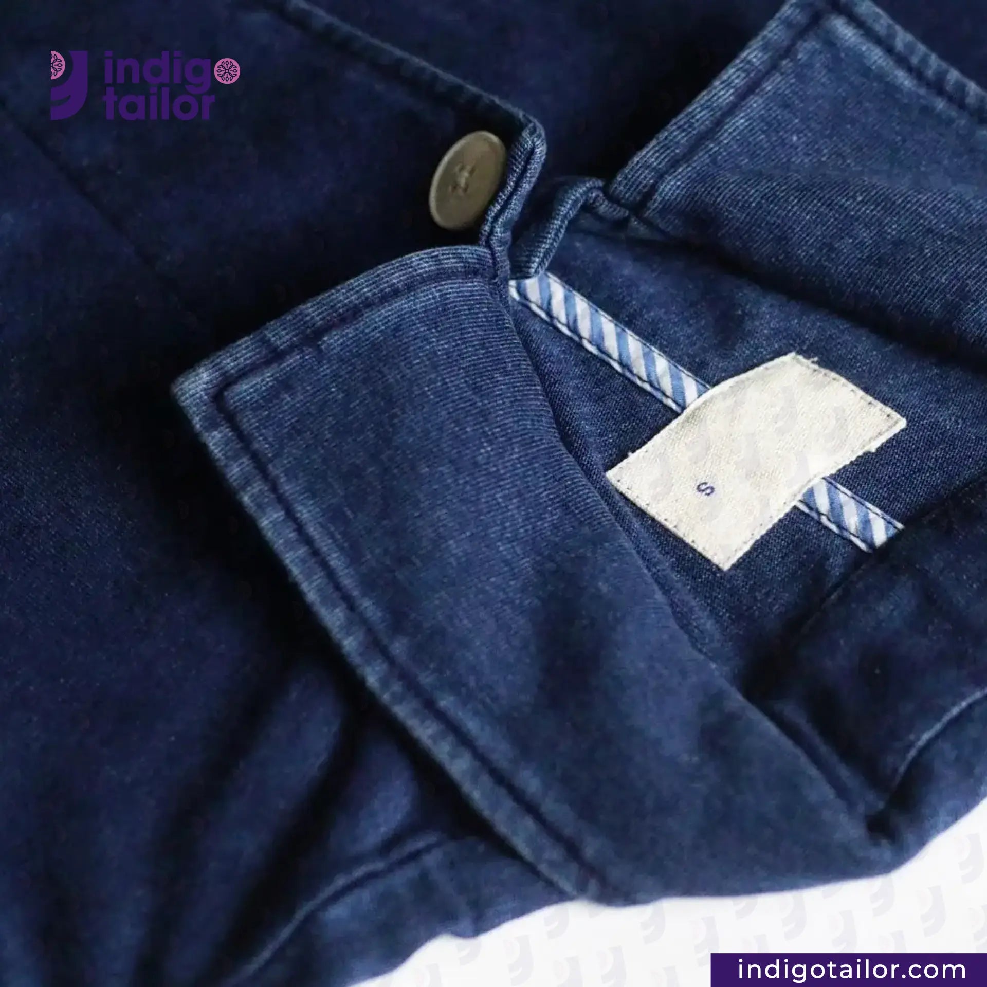 Close-up of INDIGO DYED LAB COAT with a button and label, featuring the 'Indigotailor' brand.