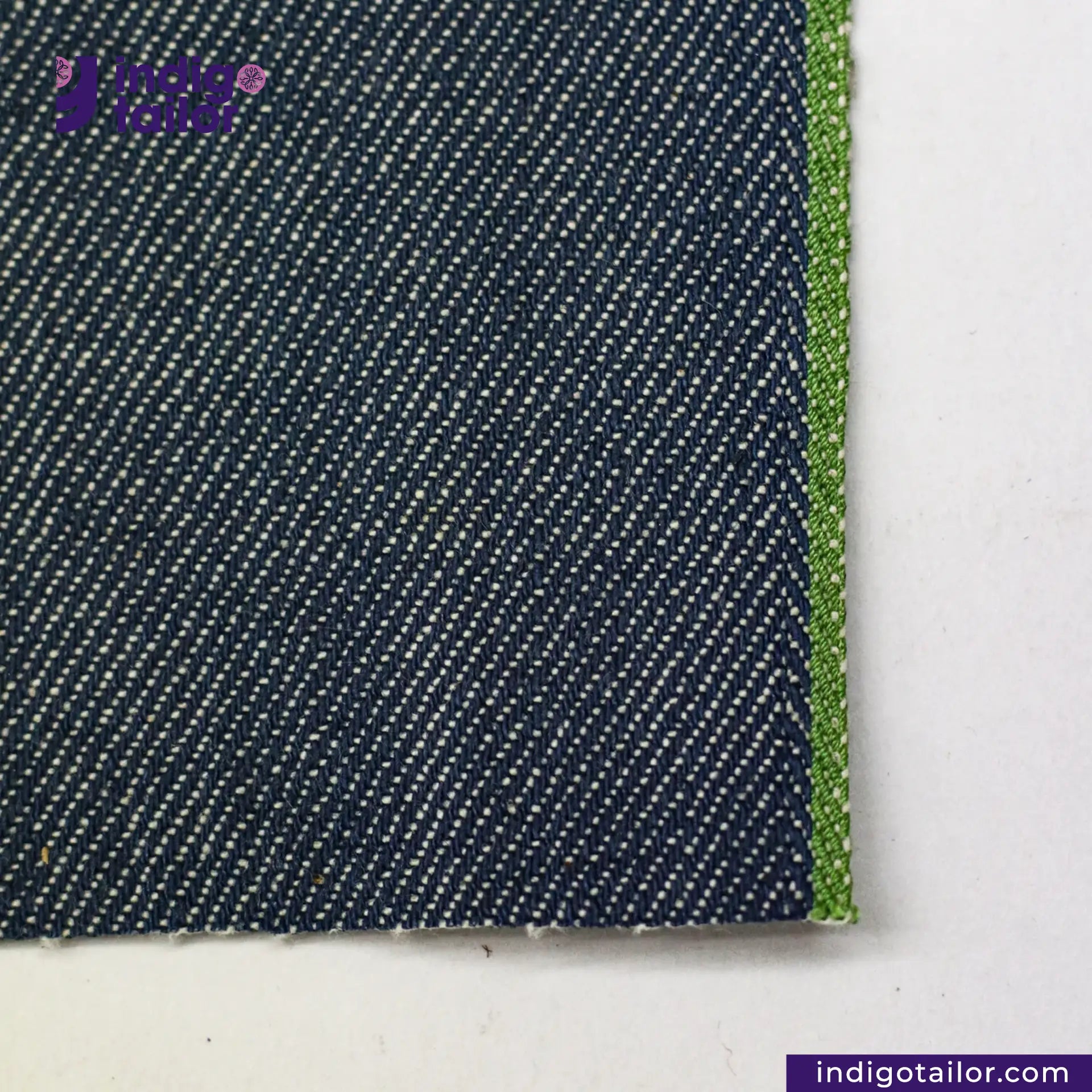 Close-up of OCEAN BLUE COTTON HEMP BLEND SELVEDGE DENIM fabric with a green edge on a white background, featuring the Indigo Tailor logo.