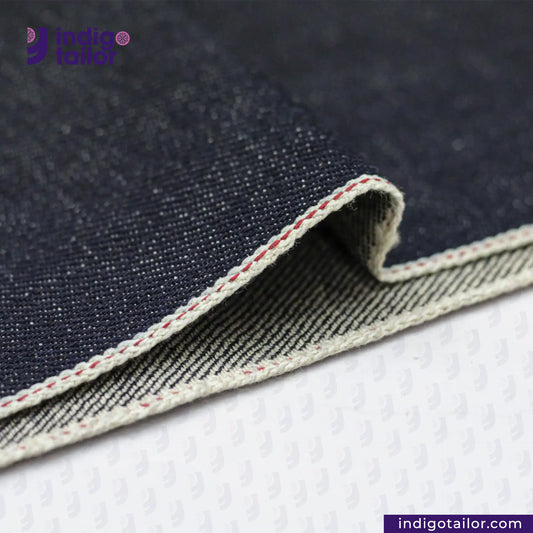 Close-up of ORGANIC NATURAL COTTON SELVEDGE DENIM fabric with visible stitching, featuring the Indigo Tailor logo.