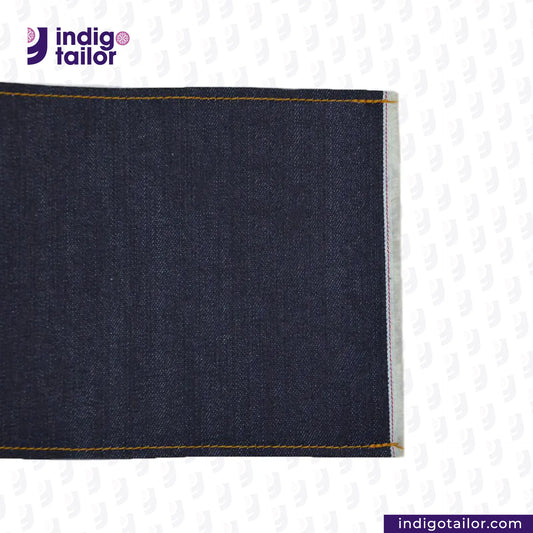 Close-up of ORIGINAL BLUE SELVEDGE DENIM fabric with yellow stitching on a light gray background, featuring the Indigotailor logo.