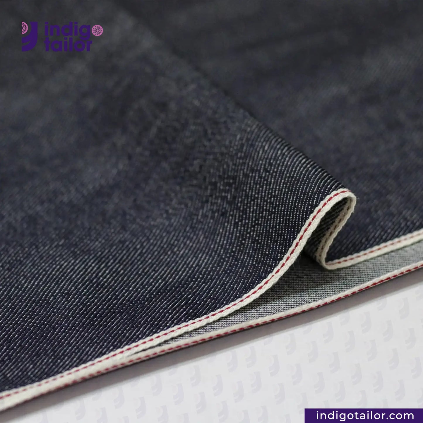 Close-up of SUPER SOFT FLUFFY SELVEDGE DENIM fabric with visible stitching on a white background, featuring the Indigotailor logo.