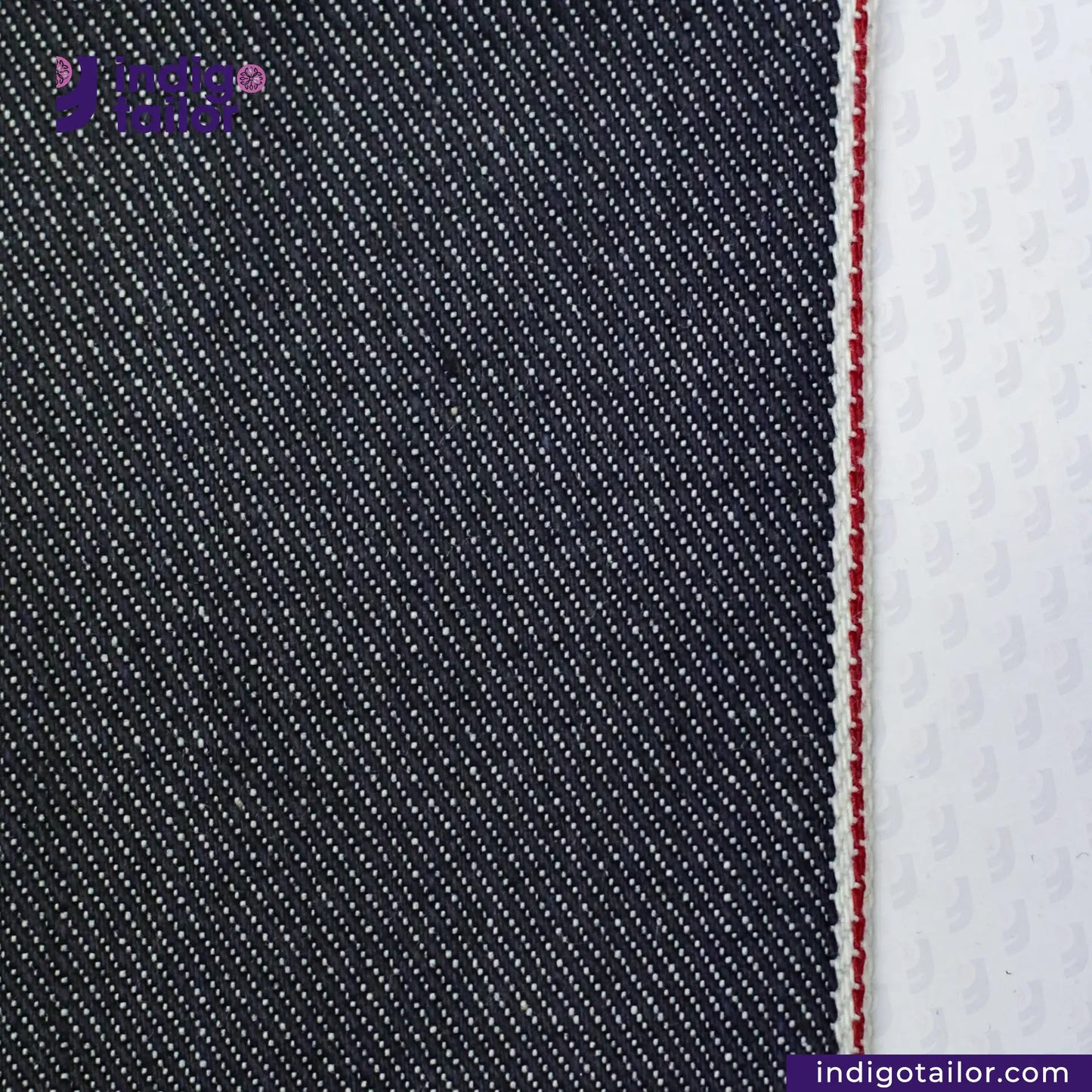 Close-up of SUPER SOFT FLUFFY SELVEDGE DENIM fabric with a white stripe and red stitching, featuring the Indigo Tailor logo.