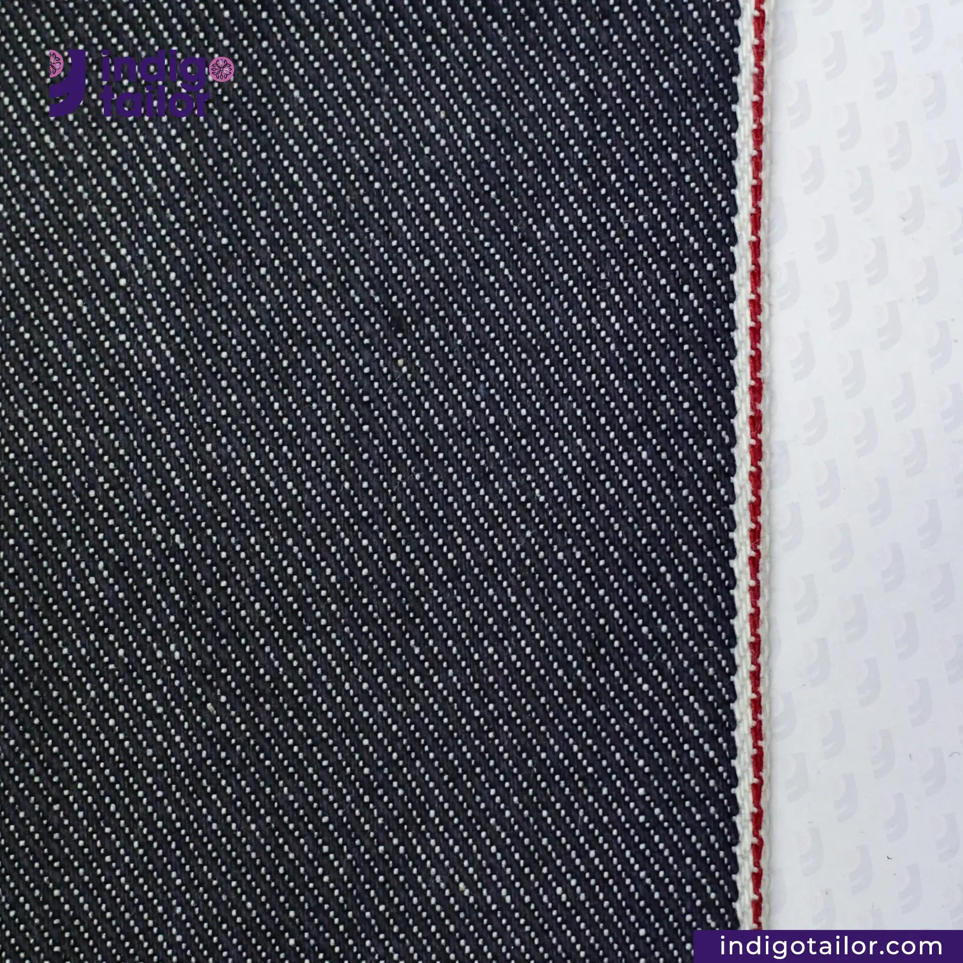 Close-up of SUPER SOFT FLUFFY SELVEDGE DENIM fabric with a white stripe and red stitching, featuring the Indigo Tailor logo.