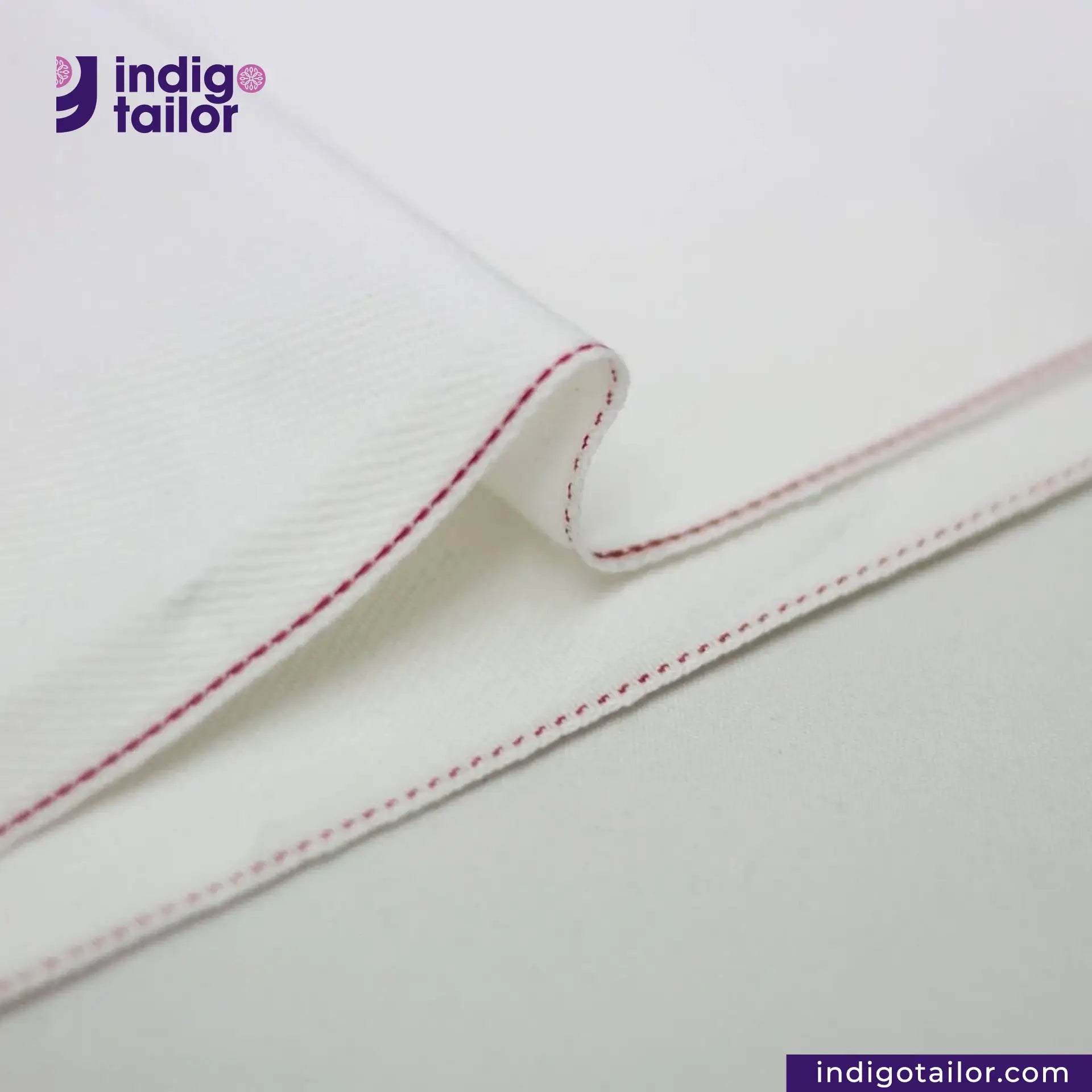 Close-up of WHITE DENIM DOTTED RED SELVEDGE fabric with red stitching on a light gray background, featuring the Indigotailor logo.